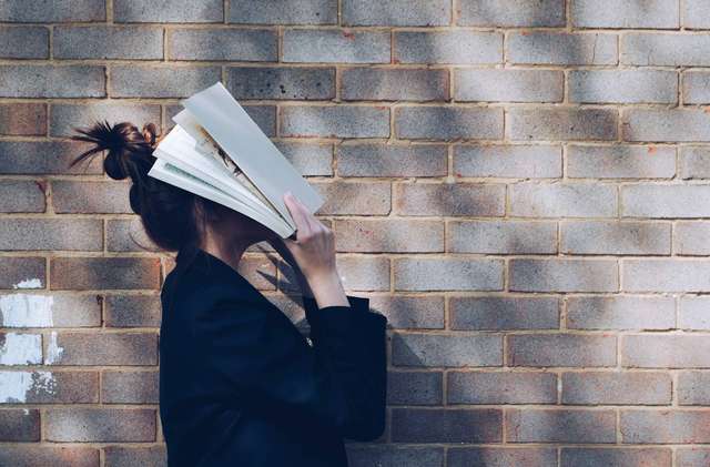 Woman standing against a brick wall with an open book covering her face.