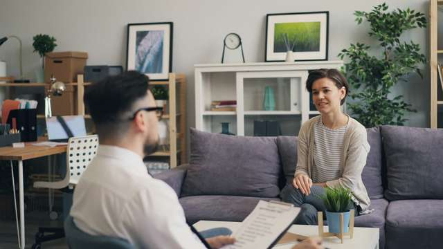 Woman sitting on a couch during a job interview with a man holding a clipboard in an office setting.