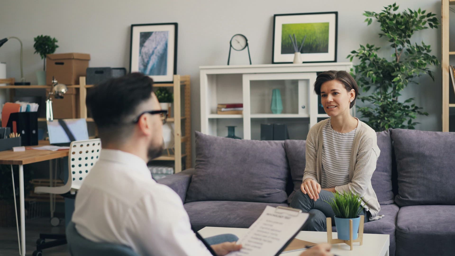 Woman sitting on a couch during a job interview with a man holding a clipboard in an office setting.