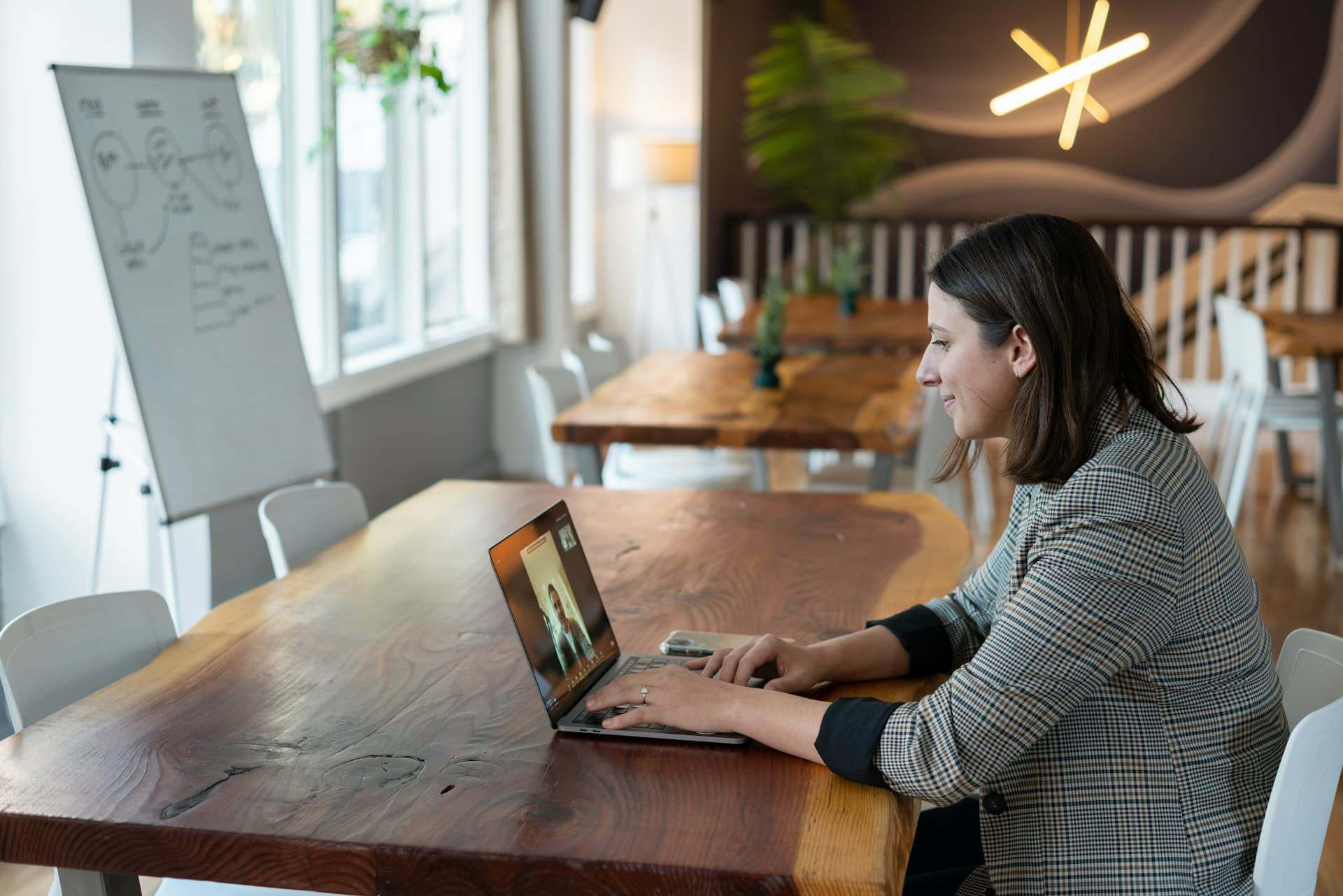 Woman in a checked blazer smiling while speaking on a virtual video call on her laptop in a modern workspace with wooden tables and a whiteboard in the background.