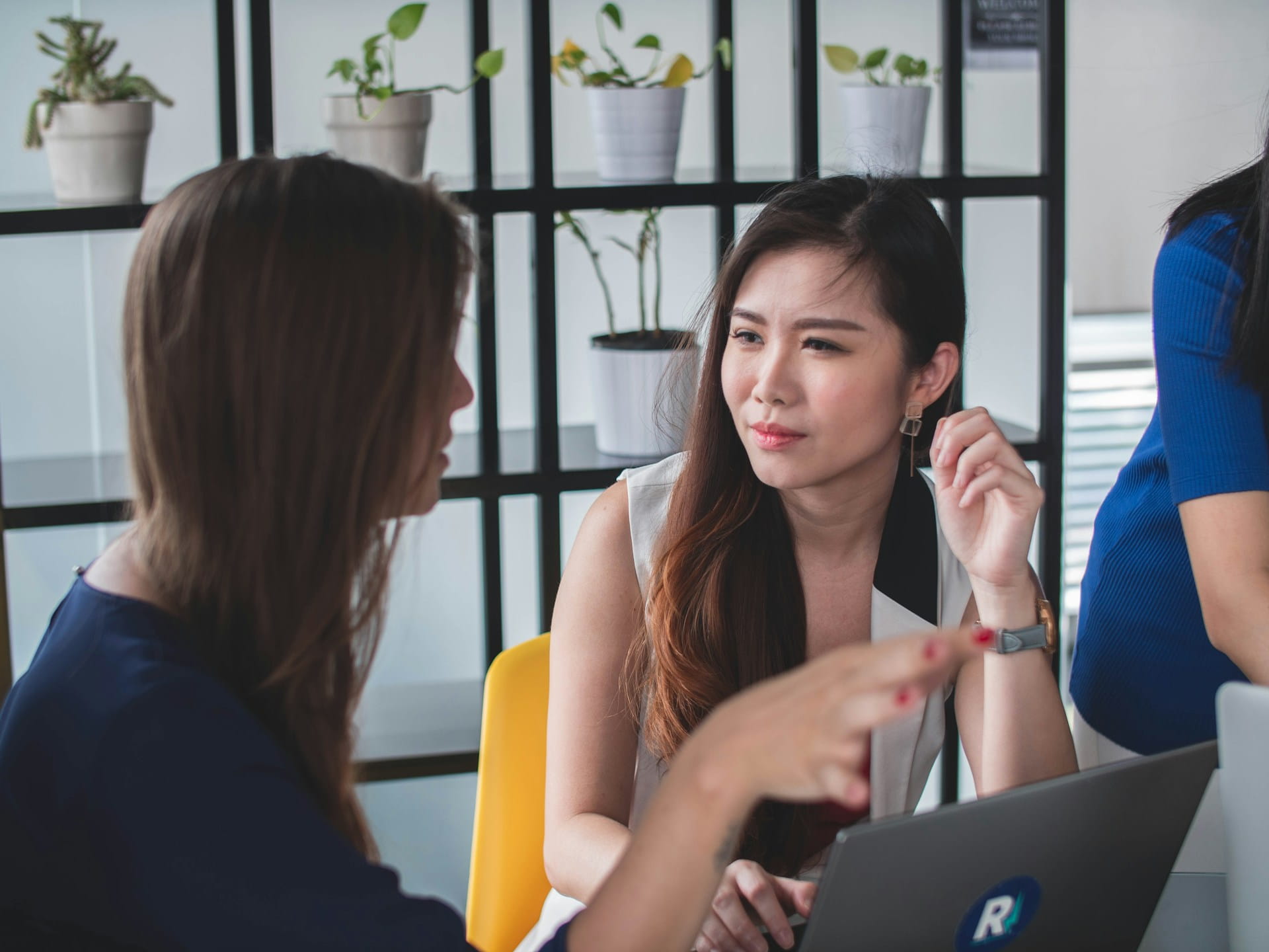 Two women engaged in a focused conversation at a desk with a laptop, in a modern office setting.
