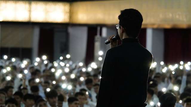 Speaker holding a microphone on stage, addressing a large audience with phone flashlights raised.