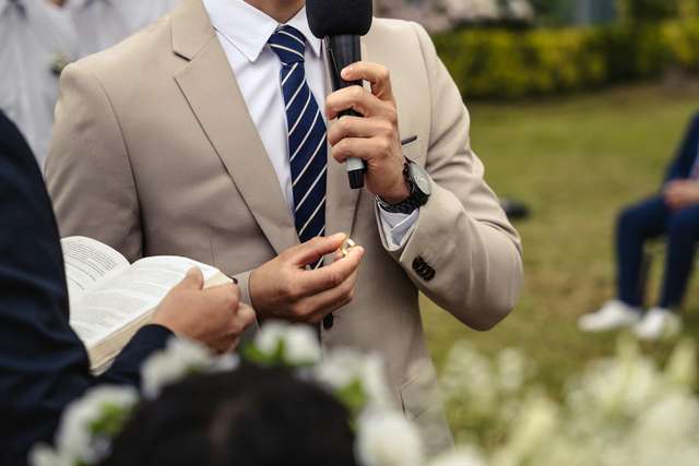 Close-up of a man in a beige suit holding a microphone and wedding rings during an outdoor ceremony.